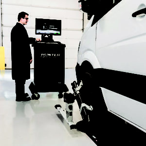 Image of the Mercedes commercial wheel aligner being used on a white medium sized van. The image is taken in a vehicle workshop with the van in the foreground and the wheel alignment machine in the background.